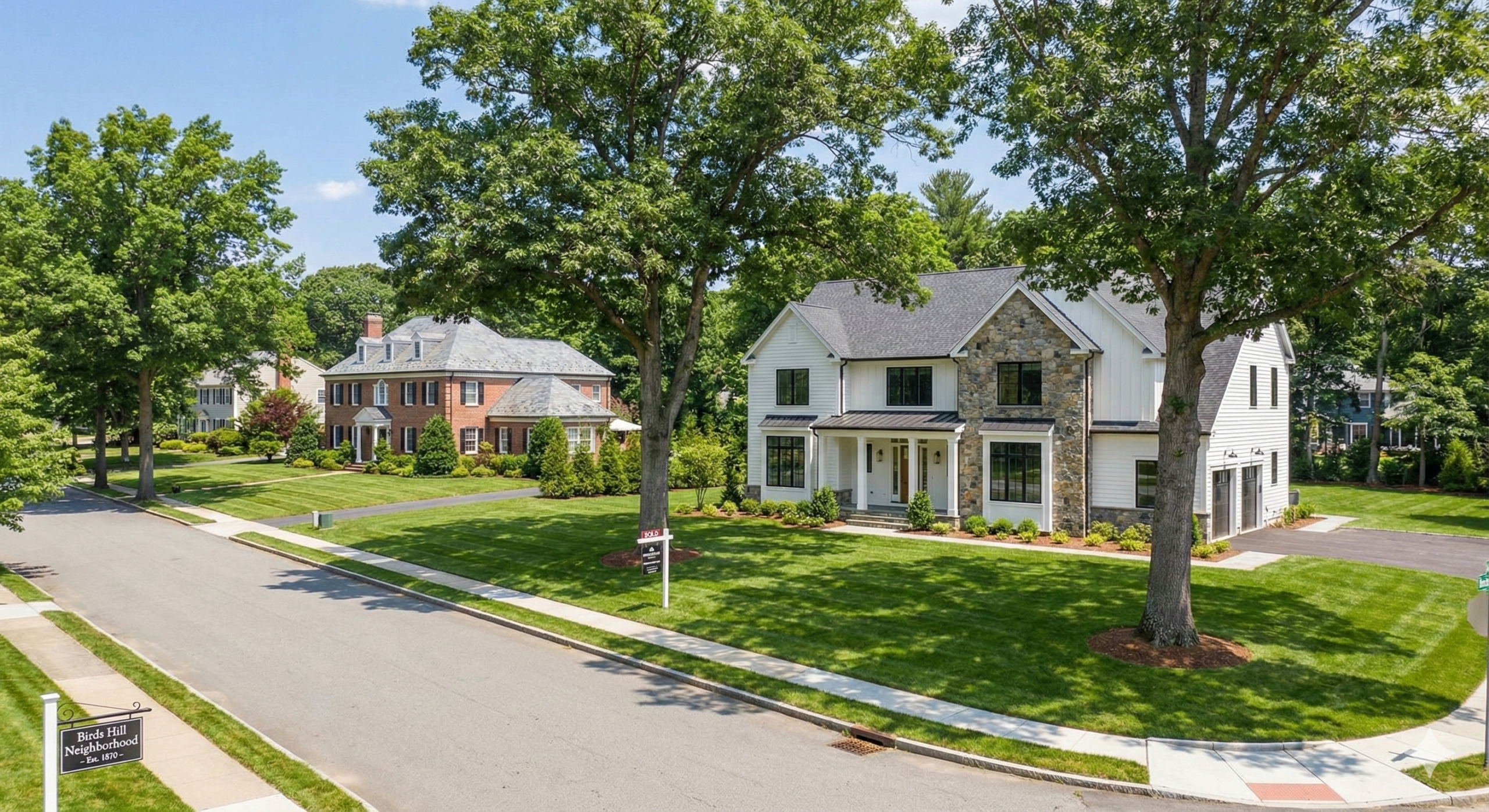 A wide-angle landscape photograph of an affluent suburban street in the Birds Hill neighborhood of Needham, Massachusetts. In the foreground, a newly constructed luxury home with a white and stone exterior and a "SOLD" sign sits on a large, manicured lawn. Across the street, a large, traditional brick colonial home is visible. The street is lined with large, mature trees, and a sign for "Birds Hill Neighborhood - Est. 1870" is in the bottom left corner.