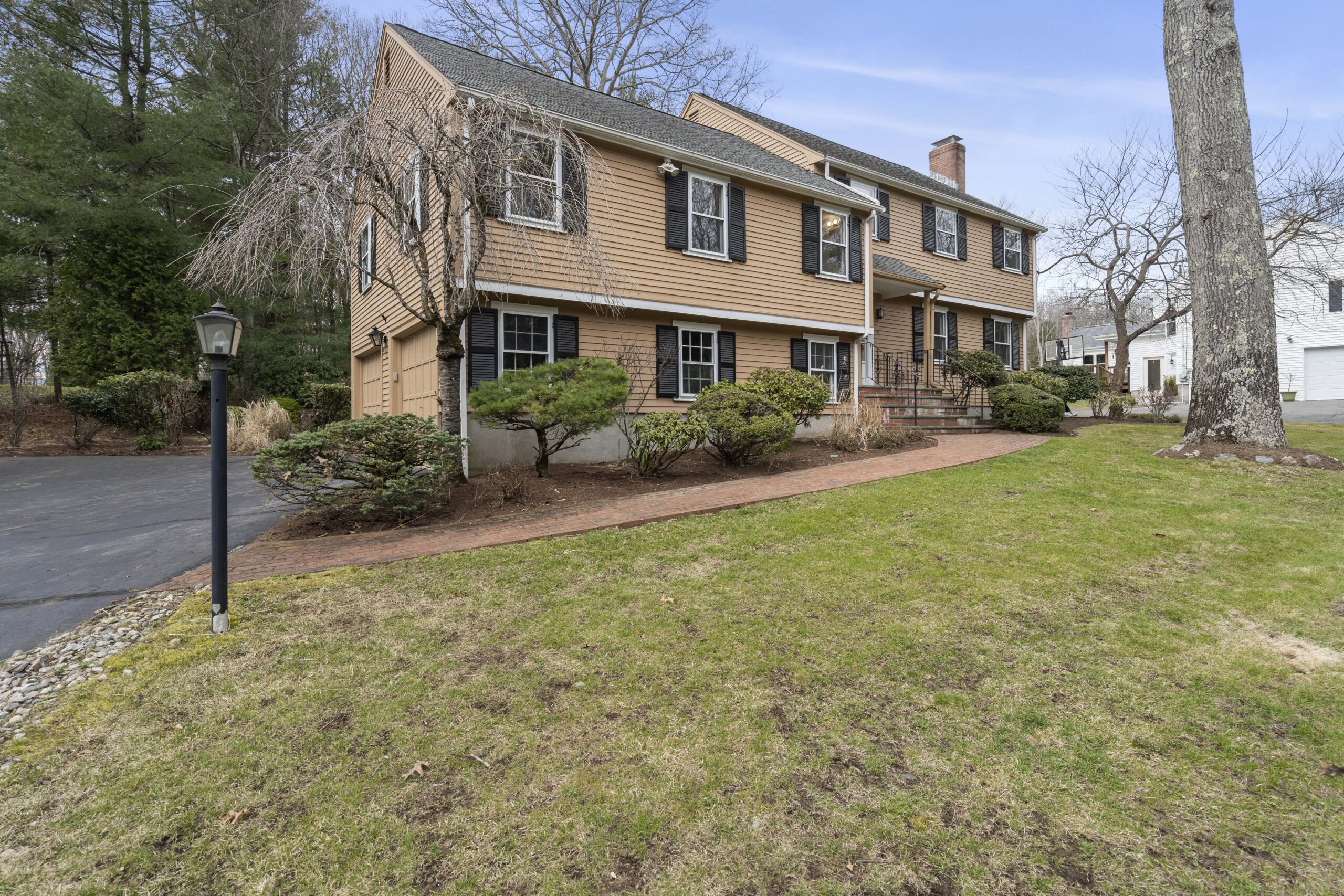 Front exterior of a two-story colonial home with tan siding, black shutters, a brick walkway and steps to the front entrance, attached garage, landscaped shrubs, and a broad front lawn with mature trees.