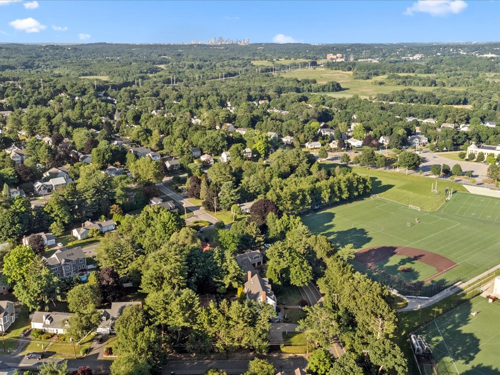 Aerial view of a tree-filled Needham neighborhood with residential streets, athletic fields, and the Boston skyline visible in the distance