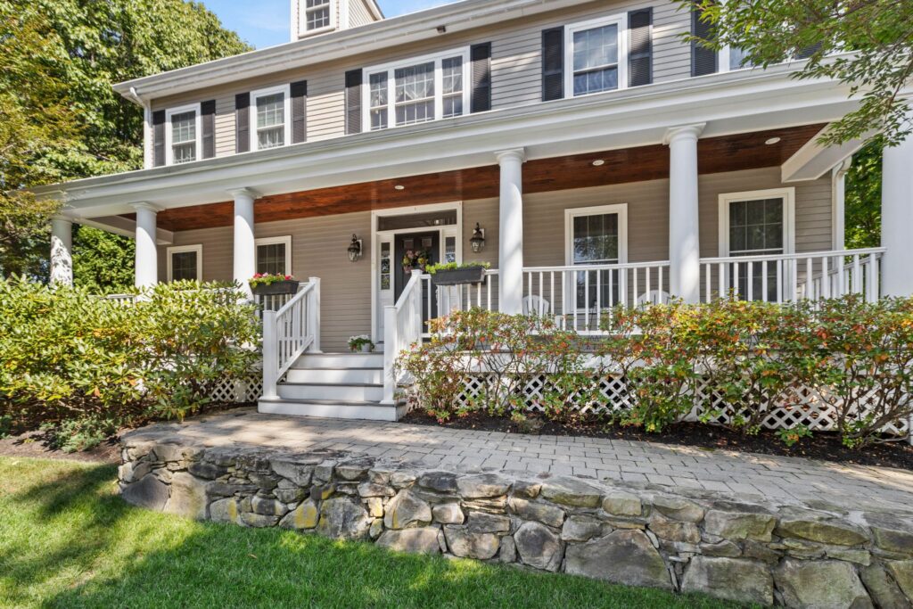 Front exterior of a gray colonial-style home with black shutters, a wide covered porch, white columns, stone retaining wall, landscaped shrubs, and mature trees.