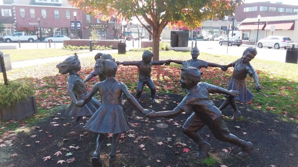 Circle of Hope sculpture in downtown Needham MA showing bronze children holding hands in a circle under a tree, with fall leaves and local storefronts in the background