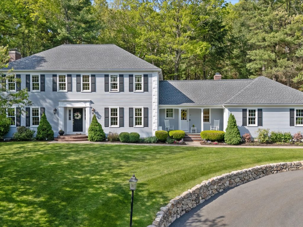 Gray Colonial-style home with black shutters, white trim, covered front entry, manicured lawn, stone wall, and circular driveway surrounded by mature trees.