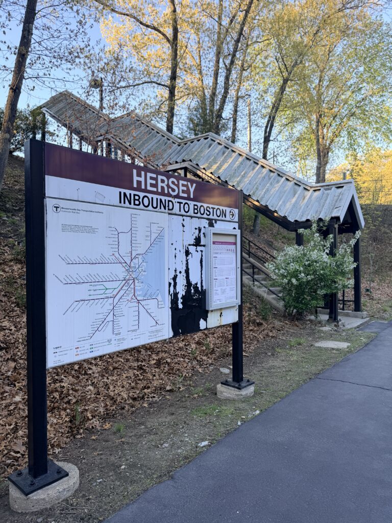 Hersey MBTA commuter rail station sign for inbound trains to Boston, with the station stairway, trees, and walking path visible in Needham, Massachusetts