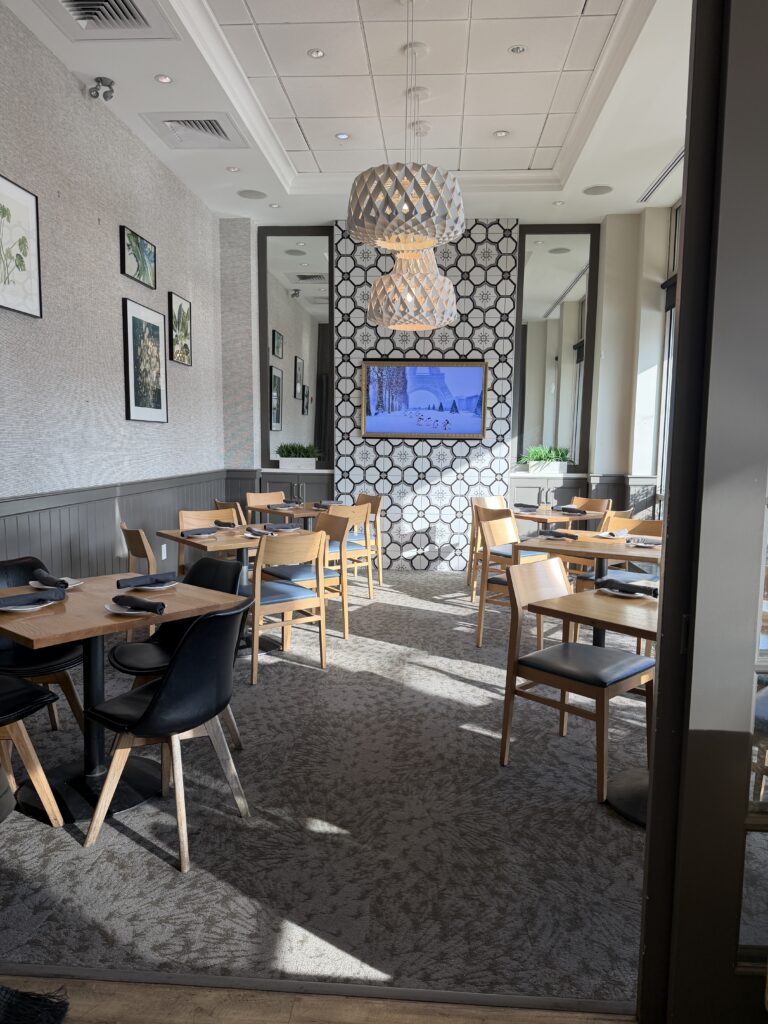 Bright restaurant dining room with wooden tables, black chairs, gray wainscoting, patterned accent wall, pendant lighting, and large windows bringing in natural light