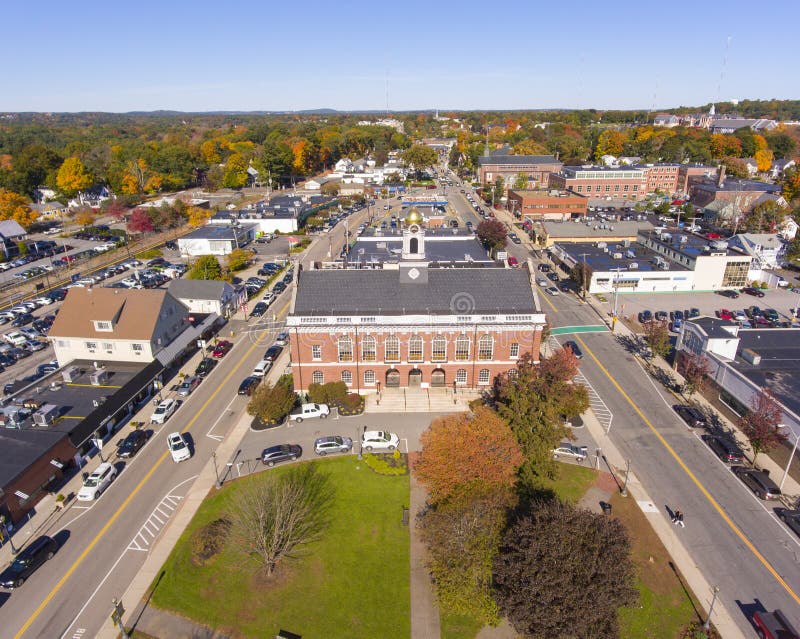 A view of downtown Needham Center, Massachusetts, showing a clean street lined with classic brick buildings, local storefronts, parked cars, and a well-maintained sidewalk under a clear blue sky.