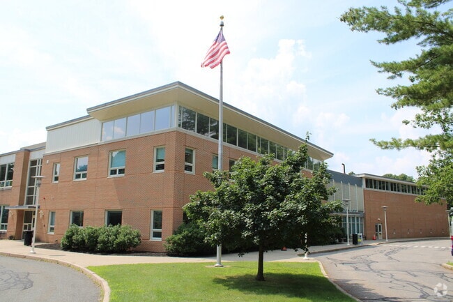 A wide-angle exterior view of the Eliot Elementary School in Needham, Massachusetts, featuring its brick facade, modern architectural design, and the school's entrance surrounded by green landscaping and a clear sky.
