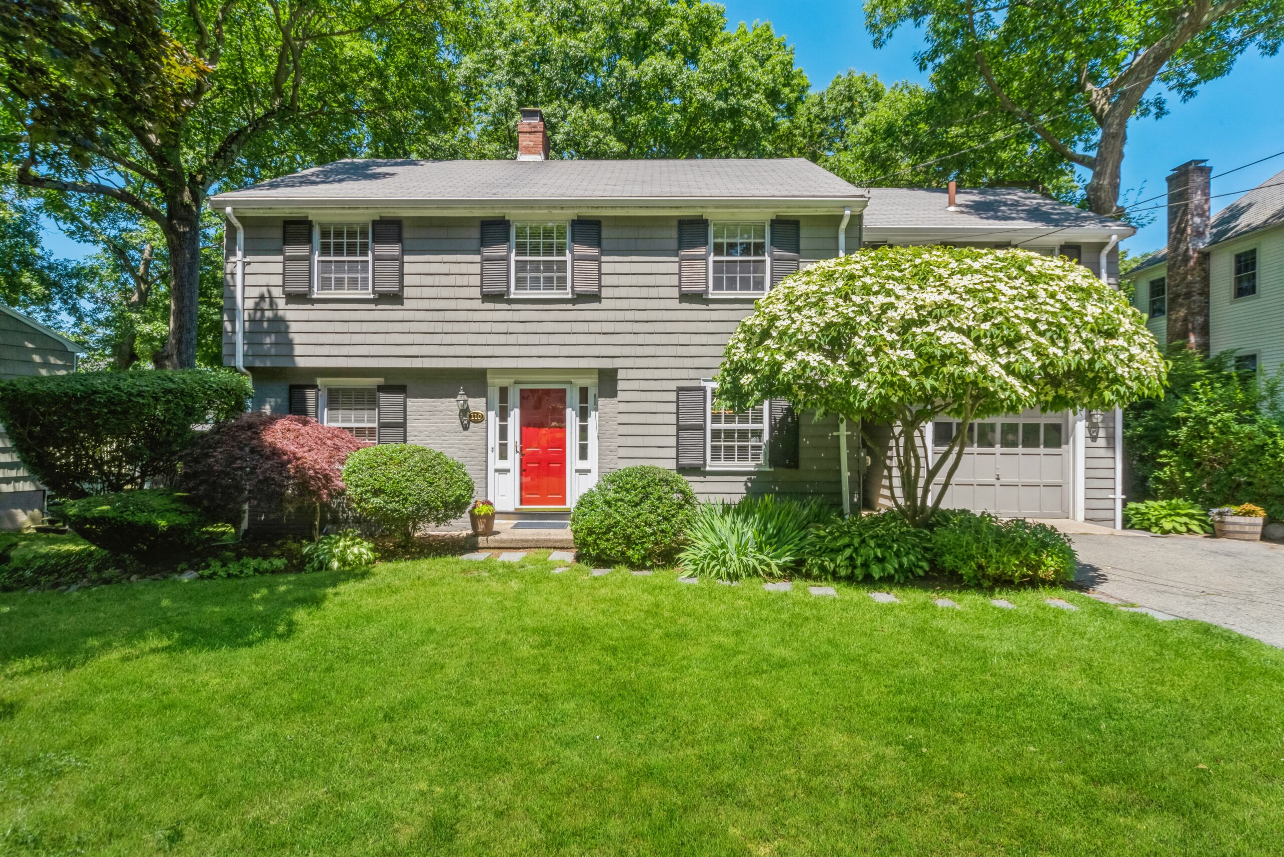 Exterior of 110 Parker Road in Needham MA featuring a gray Colonial-style home with black shutters, red front door, attached garage, mature landscaping, large front lawn, and leafy trees.