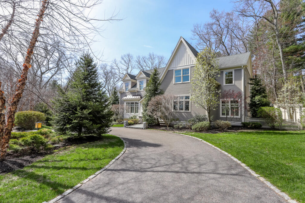 A wide-angle real estate photograph showcasing the front exterior and sweeping driveway of a large, multi-level home. The house features grey siding, white trim around multi-pane windows, and distinct steep gables, including vertical white board-and-batten siding on the prominent front peak. A paved asphalt driveway lined with stone pavers curves gently from the foreground toward the home's entrance. The property is surrounded by manicured green lawns, varied landscaping bushes, a prominent evergreen fir tree, and budding spring trees under a bright blue sky. To the far right, a small white picket fence and garden arbor are visible.