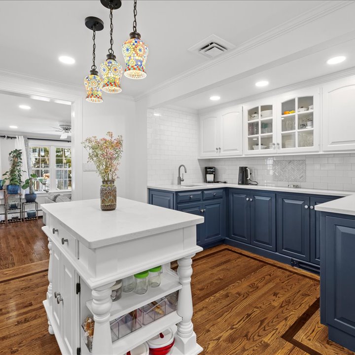 A bright, modern kitchen featuring two-toned cabinetry with white upper cabinets and deep navy blue lower cabinets. The space includes white quartz countertops, a white subway tile backsplash, and stainless steel fixtures, including a gooseneck faucet. In the foreground, a white kitchen island with a quartz top features open lower shelves containing glass jars and storage bins, topped with a vase of colorful dried flowers. Hanging directly above the island is a unique three-light pendant fixture with vibrant, mosaic-style stained glass shades. The room is finished with rich hardwood flooring that flows into an adjacent living area visible in the background.