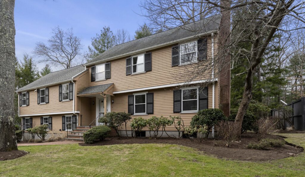 Exterior of a tan Colonial-style home at 14 Stonehurst with black shutters, brick chimney, front steps, landscaped yard, mature trees, and lawn in a quiet residential setting.