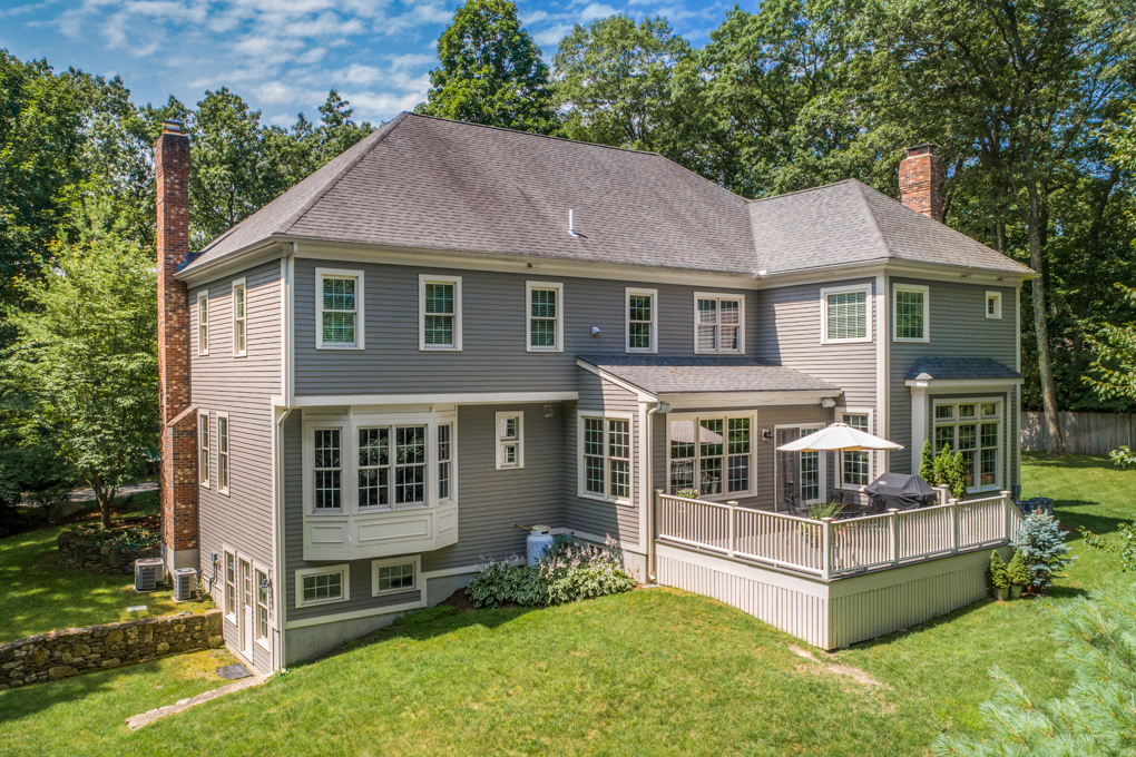 An elevated, wide-angle real estate photograph showing the spacious backyard and rear exterior of a large, two-story New England colonial-style home. The house features grey siding with white trim, a dark shingled roof, and two brick chimneys on either side. A prominent elevated wooden deck with a white railing extends from the main level, furnished with a patio umbrella and a covered grill. The property sits on a sloping green lawn surrounded by mature trees, with a small stone retaining wall visible on the lower left near a walk-out basement entrance.