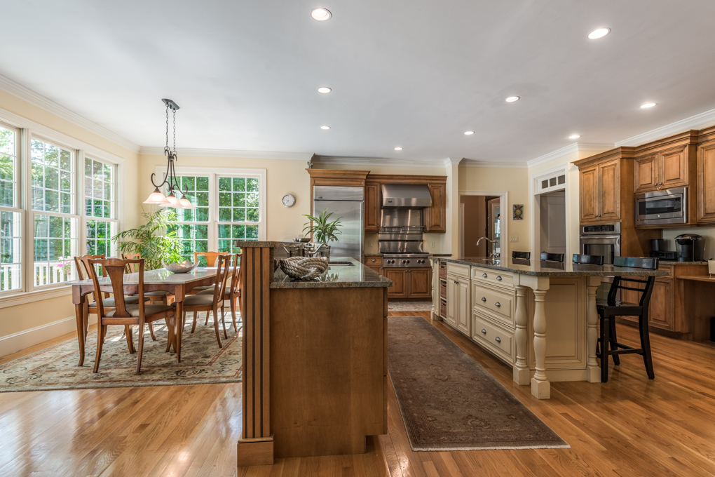 A wide-angle interior photograph of a spacious, open-concept kitchen and adjacent dining area. The kitchen features custom medium-toned wood cabinetry, stainless steel appliances—including a large professional range and built-in refrigerator—and polished granite countertops. A long, off-white kitchen island with a dark granite top and decorative turned-wood legs features built-in drawers and black barstools tucked underneath. The hardwood floor is accented by a long, dark runner rug. To the left, the sunlit breakfast area includes a dining table with a marble top, wooden chairs on a patterned area rug, and large multi-pane windows looking out to greenery.