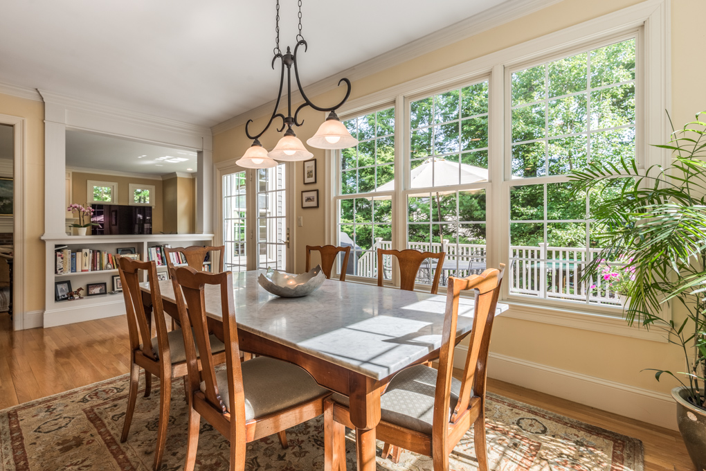 A bright, sunlit breakfast area or dining room featuring a rectangular marble-top dining table surrounded by six wooden chairs with upholstered seats. A dark metal multi-light chandelier hangs directly above the table. A large bank of three multi-pane windows fills the wall, letting in abundant daylight and offering a view of an outdoor wooden deck with a patio umbrella. The room has light yellow walls, hardwood flooring partially covered by a patterned area rug, and a half-wall with a built-in bookcase that opens up to an adjacent living area. A potted green plant sits in the right foreground.