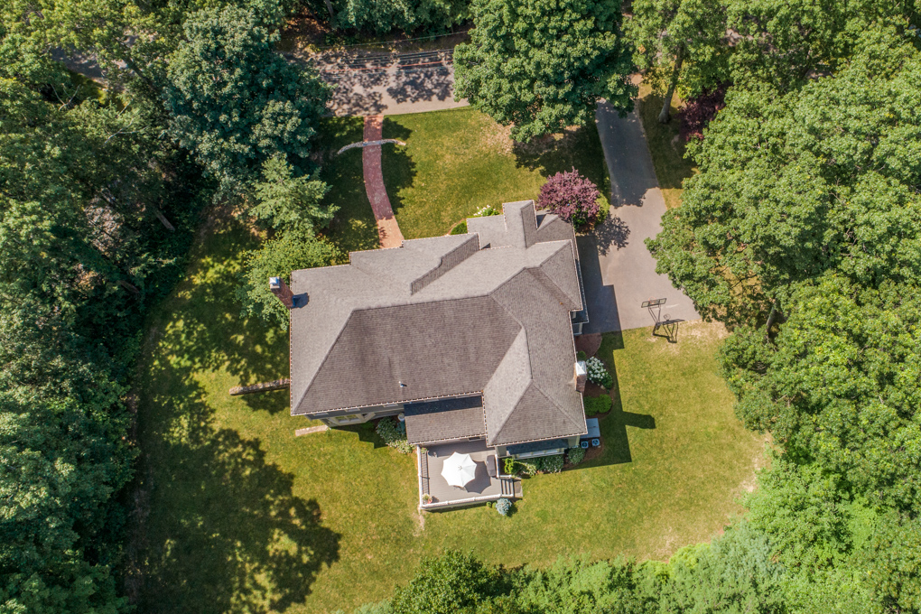 A high-angle, top-down drone photograph of a large suburban home nestled among mature, lush green trees. The complex grey shingled roofline of the multi-level house dominates the center of the frame. Below the house, a wooden back deck with a white patio umbrella faces a spacious grassy backyard. A paved driveway sits on the right side of the property featuring a basketball hoop, while a brick walkway cuts through the front lawn toward a street visible at the top of the image. Tree shadows stretch across the well-maintained lawns.