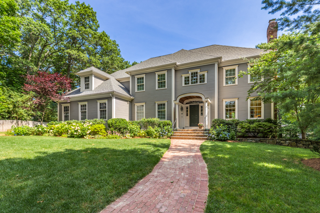 A wide-angle real estate photograph showcasing the grand front exterior of a large, multi-level colonial-style home. The house features grey horizontal siding, crisp white trim around numerous multi-pane windows, and a dark shingled roof. A curved red brick walkway cuts through a vibrant, sloping green front lawn, leading directly to a small brick entryway staircase and an arched front portico protecting a dark front door. Lush green bushes, manicured landscaping, and flowering hydrangeas line the foundation of the house, which is surrounded by large, mature green trees under a clear blue sky.