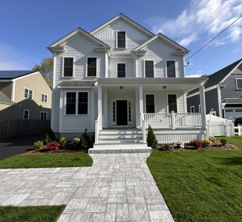 White new construction home at 303 Brookline Street in Needham MA with covered front porch, stone walkway, landscaped front yard, and bright blue sky.