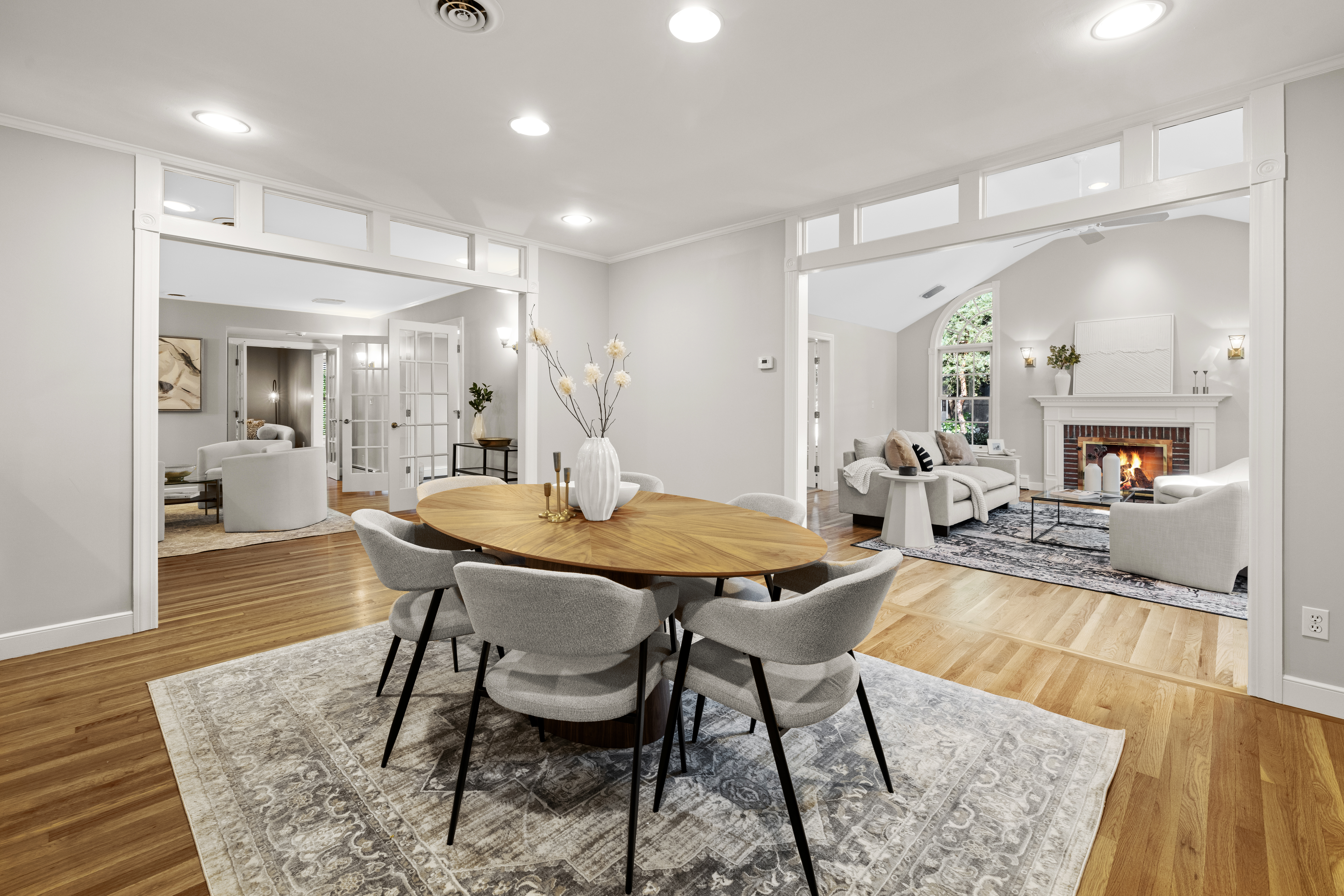 Bright staged dining room at 45 South Street in Needham MA with hardwood floors, round wood dining table, neutral chairs, transom windows, and open views into adjoining living and sitting rooms with fireplace.
