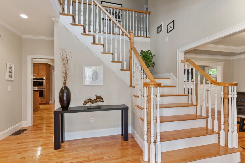 A wide-angle interior photograph of a spacious, well-lit home foyer featuring a central wooden staircase. The staircase has oak treads, white risers, white spindles, and a matching oak handrail that curves at the landing. To the left of the stairs, a narrow black console table is decorated with a tall dark vase containing dried branches and a piece of driftwood art. The walls are painted a light neutral beige with crisp white trim, and polished hardwood floors run throughout the space. In the background, doorways open up to reveal a kitchen with wooden cabinetry on the left and a separate living area on the right.