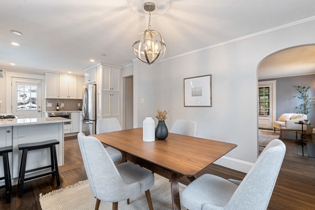 A bright, wide-angle interior photograph of an open-concept dining room and kitchen area. In the foreground, a rectangular wooden dining table is surrounded by four light grey upholstered chairs, sitting on a textured woven rug over dark hardwood floors. A spherical gold and silver metal chandelier hangs over the table, which is decorated with a white vase and a black vase with dried pampas grass. The background features a kitchen with white cabinetry, a marble-topped island with black barstools, and stainless steel appliances. To the right, a large arched doorway opens into an adjacent living room where a white armchair and side table are visible.