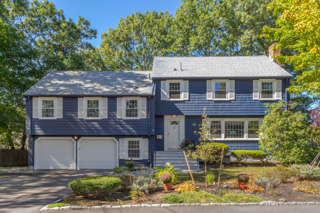 Exterior of a blue split-level home on Aletha Road in Needham MA with white shutters, two-car garage, landscaped front yard, mature trees, and sunny residential setting.