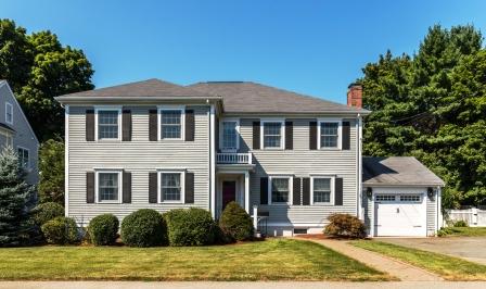 Exterior of a gray Colonial-style home on Bird Street in Needham MA with black shutters, attached garage, front lawn, mature trees, and a sunny residential setting.