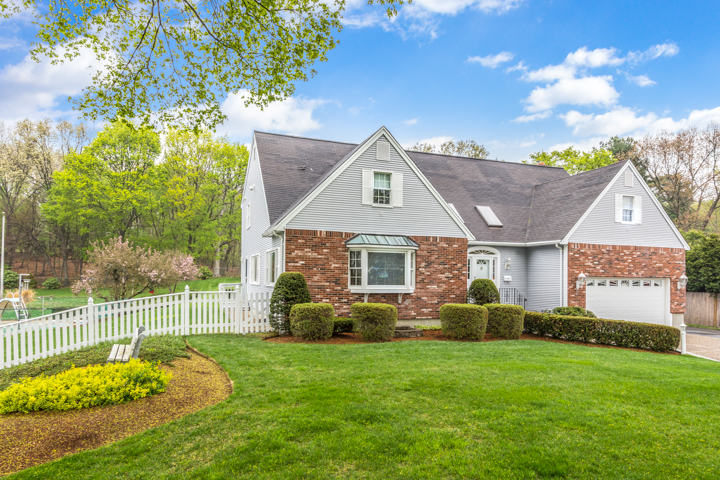 Exterior of a brick and gray shingle home on Hunnewell Street in Needham MA with a manicured front lawn, white picket fence, attached garage, and landscaped yard under a bright blue sky.