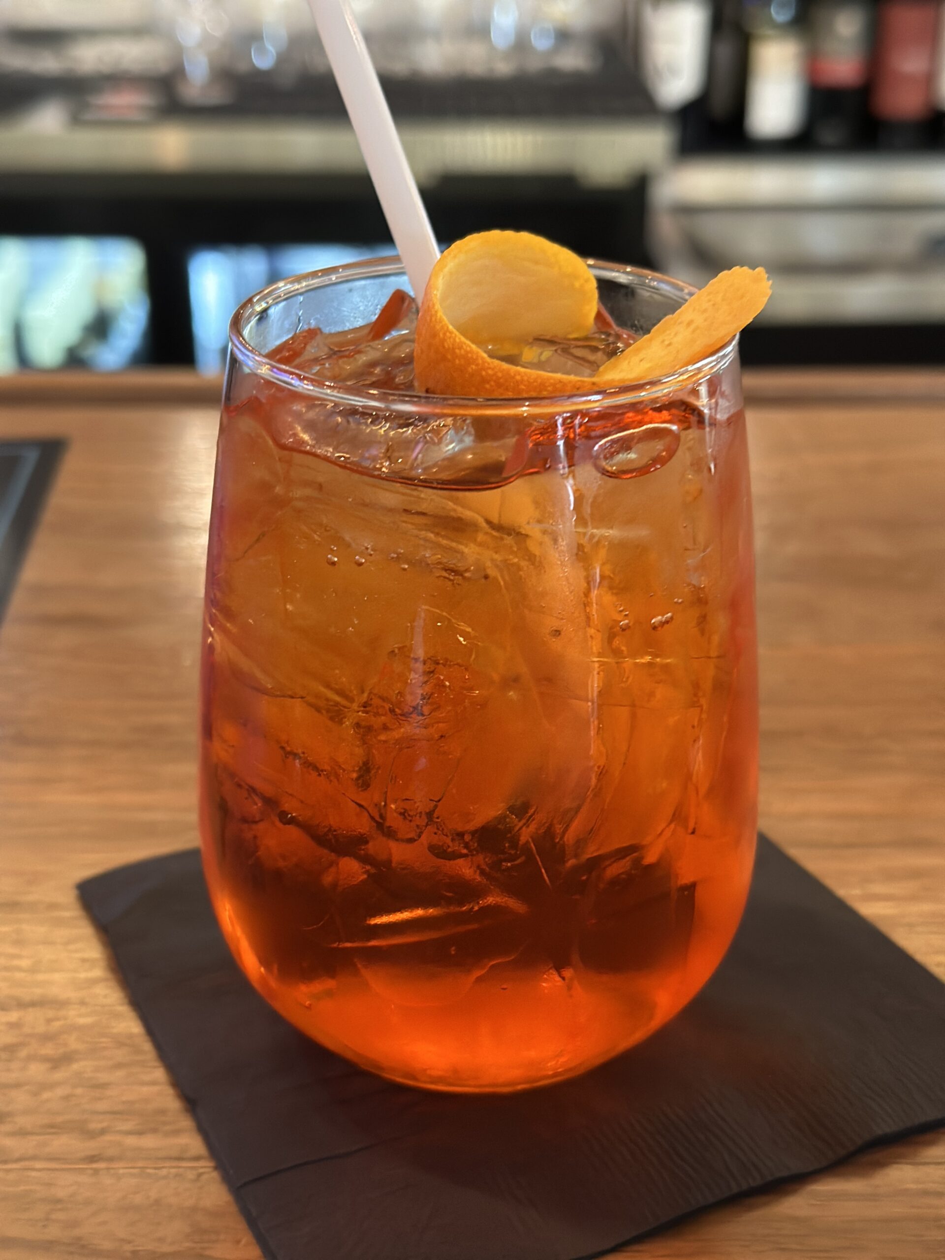 A close-up, eye-level photograph of a vibrant orange cocktail, likely an Aperol Spritz, served in a stemless wine glass filled with ice cubes. The drink is resting on a black square napkin on a smooth wooden bar top. It is garnished with a wide, twisted orange peel resting across the rim and has a white plastic straw sticking out. The background features a softly blurred, out-of-focus view of a bar area with bottles on display shelves.