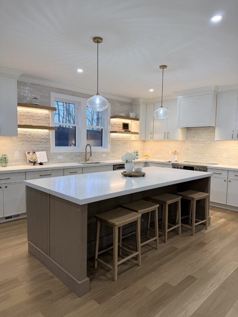 Modern luxury kitchen with a large waterfall-edge island, three wood counter stools, white cabinetry, warm under-cabinet lighting, glass pendant lights, wide-plank wood floors, and a white subway tile backsplash.