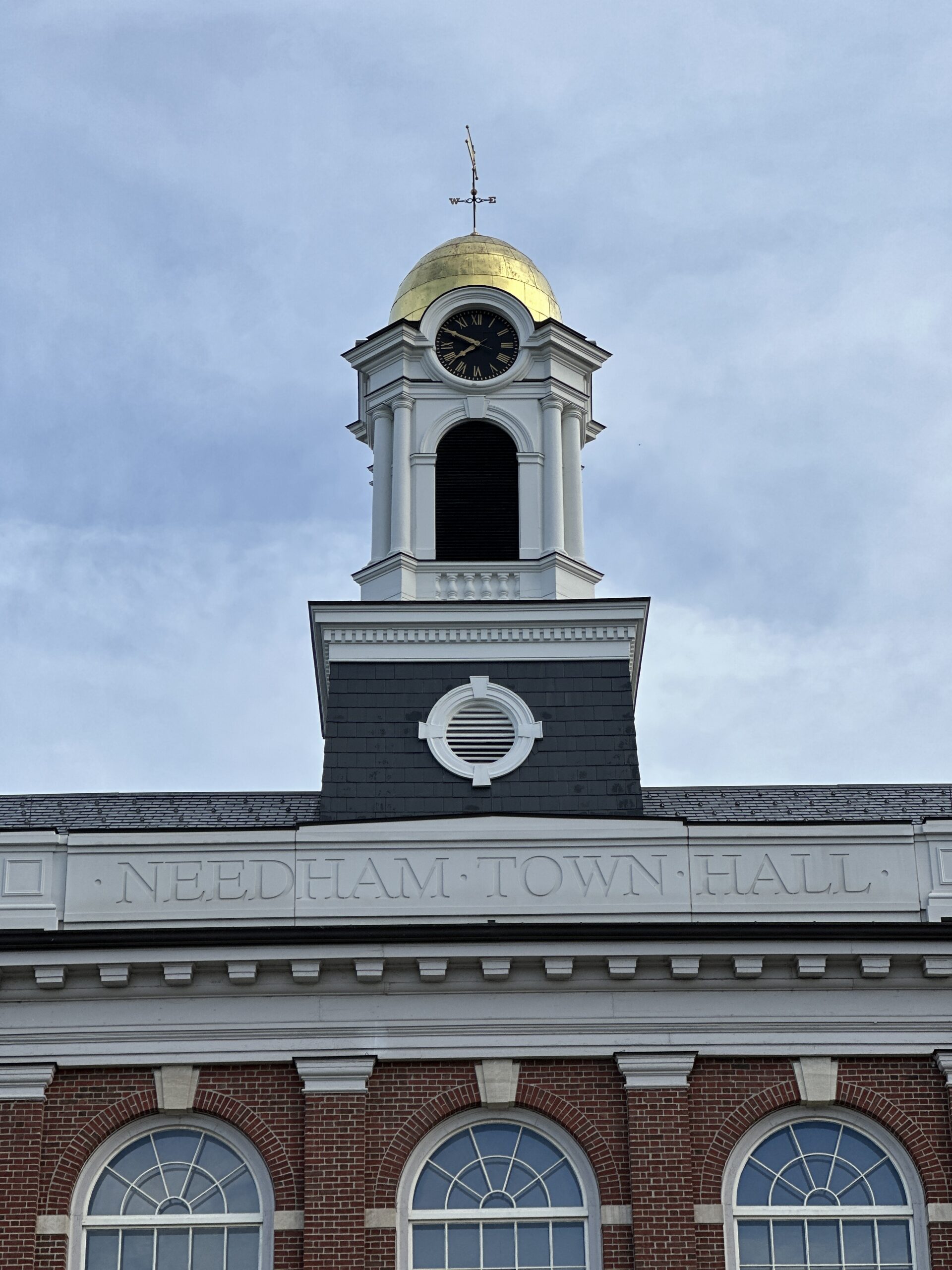 Close-up of Needham Town Hall in Needham MA showing the historic brick facade, arched windows, clock tower, gold dome, and weathervane against a cloudy sky.