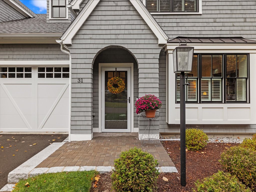 Gray shingle-style home in Needham, MA, featuring an arched covered entry, white garage door, brick walkway, landscaped front garden, and a welcoming wreath on the front door.