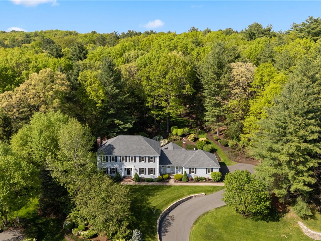 Aerial view of a classic Colonial-style home in Needham, MA, surrounded by mature trees, landscaped grounds, and a curved driveway.