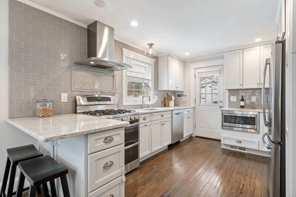 Bright updated kitchen in Needham, MA, featuring white cabinetry, marble countertops, gray subway tile, stainless-steel appliances, breakfast-bar seating, and hardwood floors.