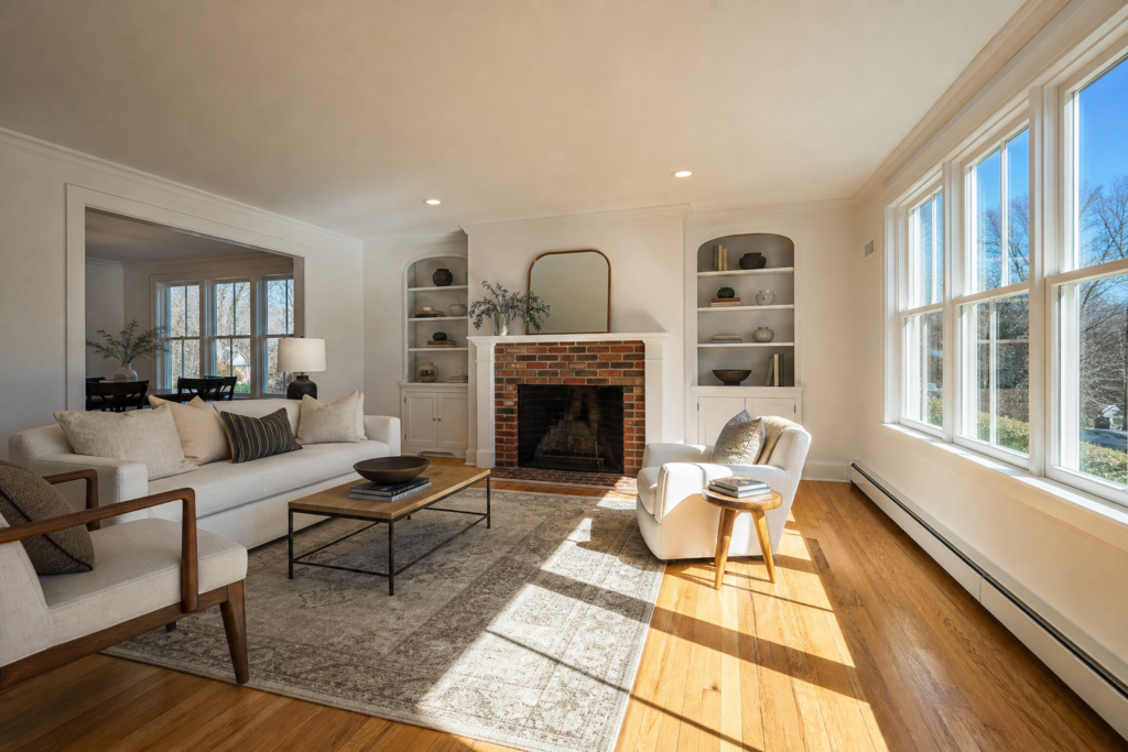 Sunlit living room with brick fireplace, built-in bookshelves, and large windows overlooking a wooded lot