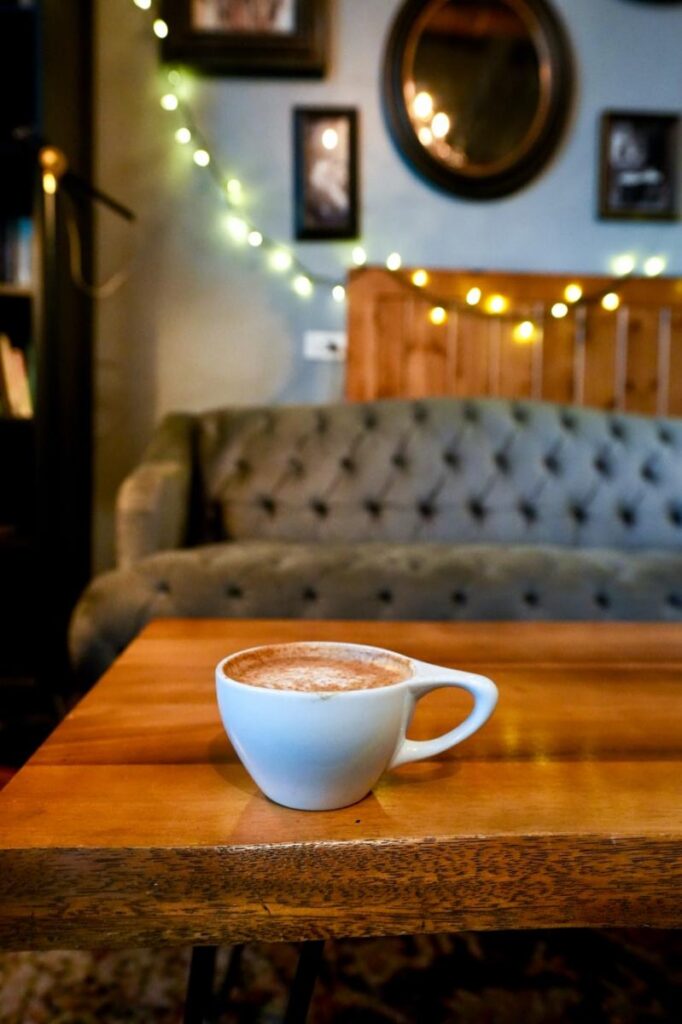 White cappuccino cup on a wooden coffee table inside a cozy coffee shop in Medfield MA, with a tufted velvet sofa and string lights in the background