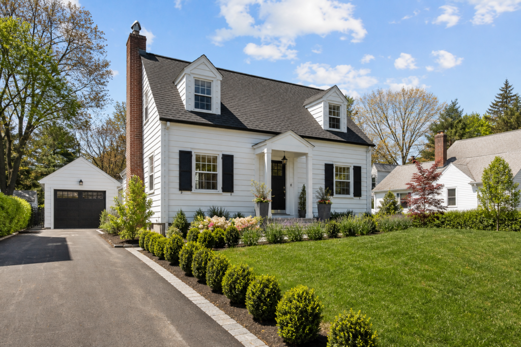 Bright exterior view of a charming white Cape-style home with black shutters, dormer windows, a detached one-car garage, manicured landscaping, and a green front lawn under a blue sky.