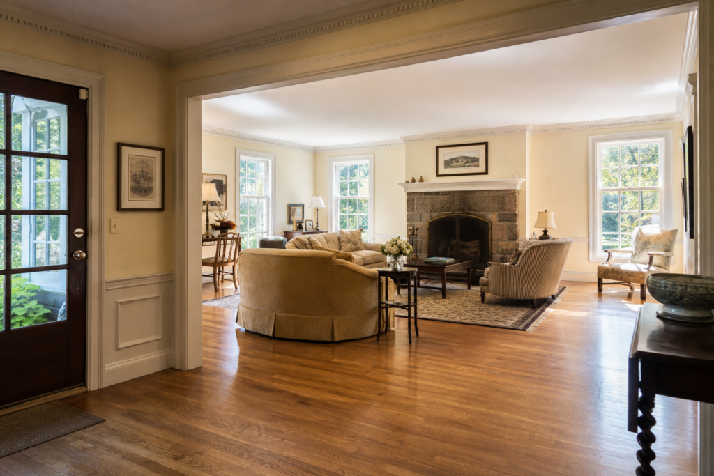 Spacious traditional living room with hardwood floors, a large stone fireplace, neutral furnishings, abundant natural light, and detailed crown molding.