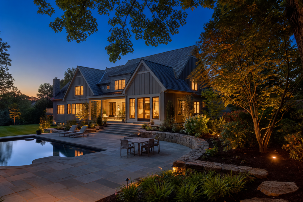 Luxury home at twilight with warmly lit windows, a stone patio, swimming pool, lounge chairs, outdoor dining area, and professionally illuminated landscaping.