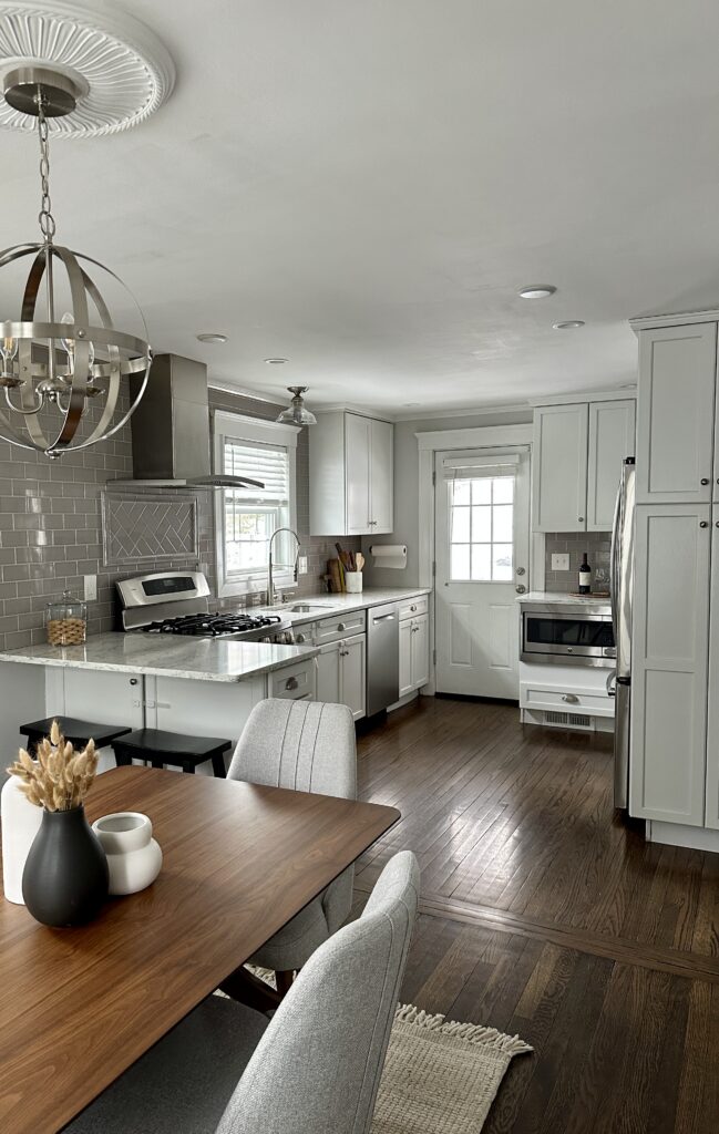 Updated kitchen and dining area in Needham, MA, featuring light gray cabinetry, marble countertops, stainless-steel appliances, subway tile backsplash, dark hardwood floors, and a wood dining table.