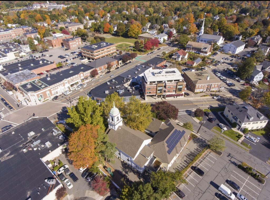 Aerial view of Needham Center, MA, showing local shops, streets, churches, parking areas, and colorful fall foliage.