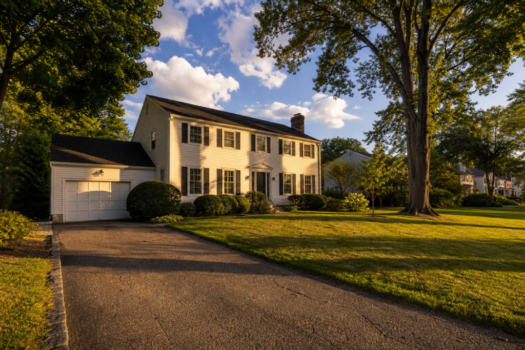 White two-story Colonial home in Needham, Massachusetts, with black shutters, an attached garage, manicured landscaping, and a spacious lawn shaded by mature trees in warm evening light.