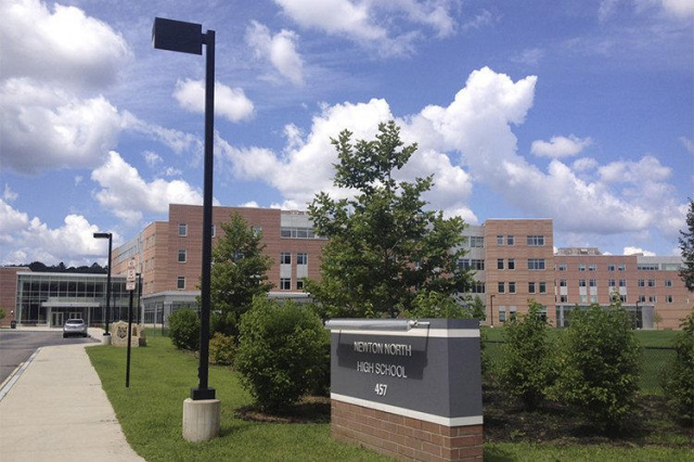 Exterior of Newton North High School featuring a large brick campus, landscaped entrance, sidewalk, and school sign beneath a partly cloudy sky.