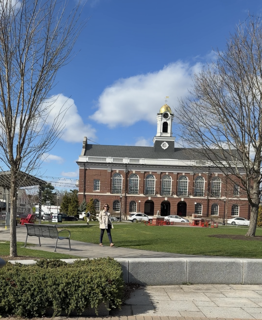 Needham Town Hall with its gold-domed clock tower behind the town common, framed by bare trees, blue sky, and a woman walking along the pathway.