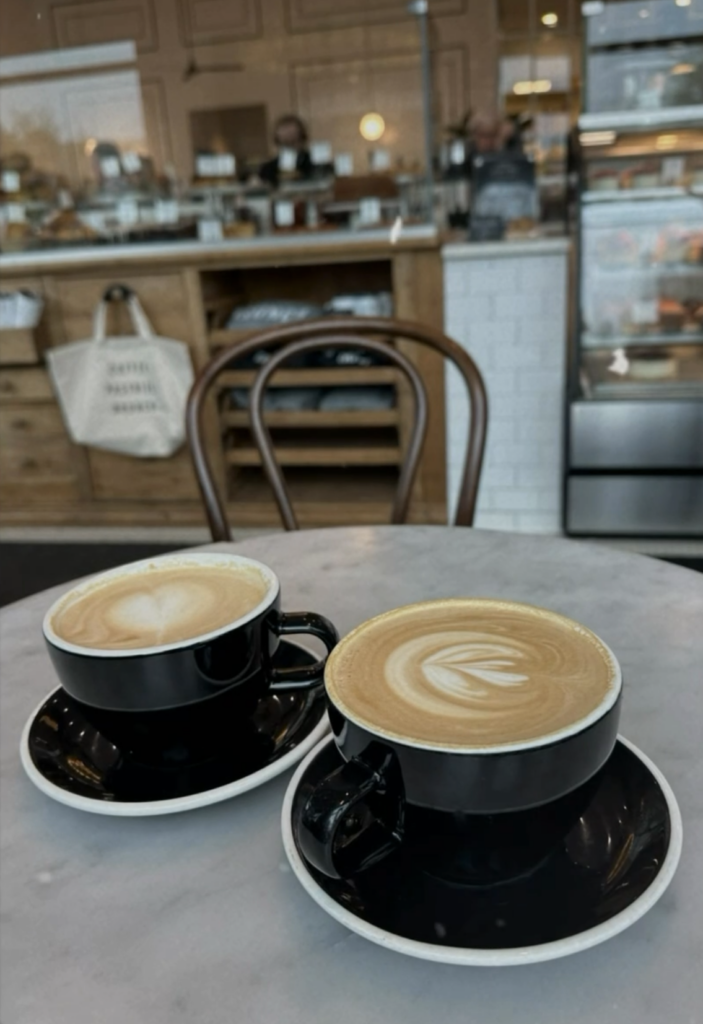 Two latte art coffees on black cups and saucers at a marble café table, with a bakery counter in the background