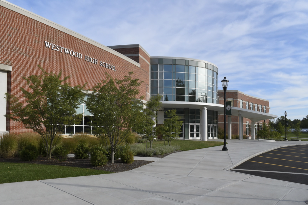 Exterior of Westwood High School featuring a red-brick façade, curved glass entrance, landscaped walkway, and blue sky.