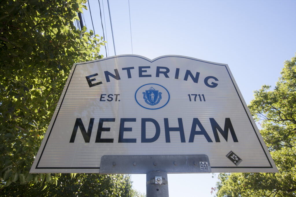 Entering Needham town sign featuring the Massachusetts state seal and the town’s 1711 founding date, surrounded by leafy trees under a clear blue sky.