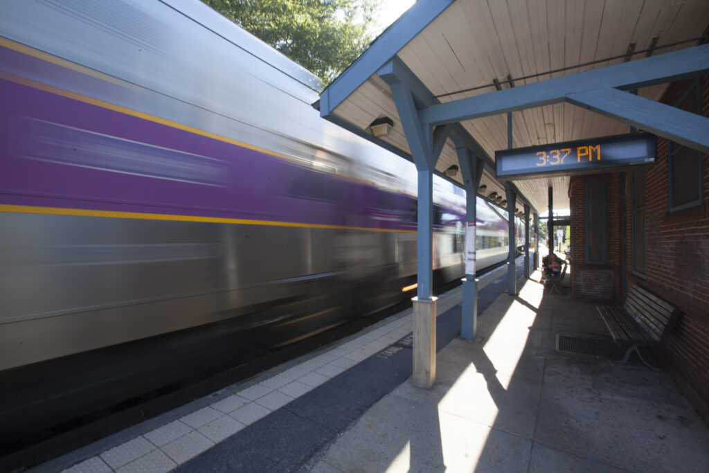 MBTA Commuter Rail train passing through Needham Junction station in Needham, MA, beside a covered platform and digital clock.
