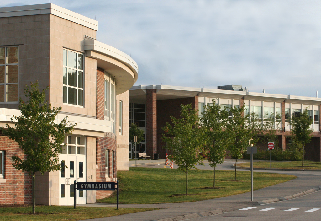 Exterior of Medfield High School featuring a brick-and-stone façade, gymnasium entrance, landscaped lawn, trees, and a curved driveway.