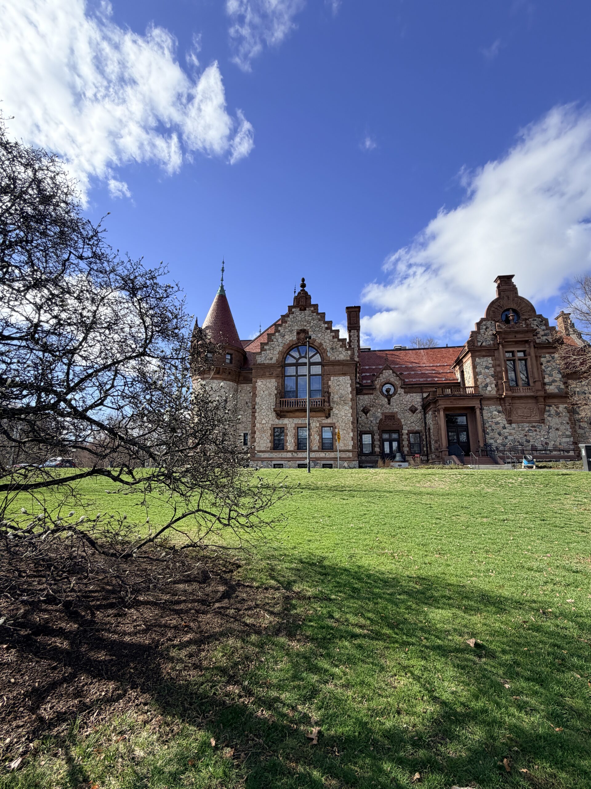 Historic town hall stone building in Wellesley, Massachusetts, featuring arched windows, decorative brickwork, and a turret overlooking a green lawn beneath a bright blue sky.