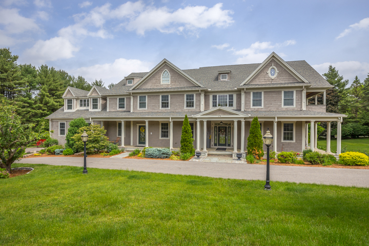 Front exterior of a large gray-shingled home in Needham, MA, with white trim, a covered porch, circular driveway, landscaped grounds, and expansive lawn