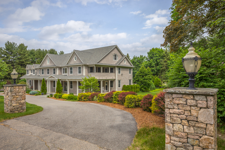 Side-front view of a large gray-shingled home in Needham, MA, with a circular driveway, stone pillars with lanterns, covered porches, and colorful landscaped gardens